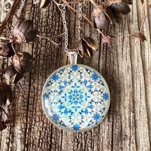 May include: A silver pendant necklace with a round, patterned pendant. The pendant features a blue and white geometric design. The necklace hangs against a rustic wooden background, with dried seed pods in the upper portion of the image.