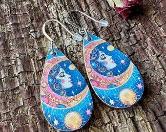 May include: A pair of teardrop-shaped earrings with a celestial design. The earrings feature a woman's face with a crescent moon and stars on a blue background. The earrings are made of wood and have silver hooks.