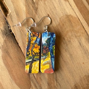 May include: A pair of silver earrings with rectangular wood panels featuring a painting of a landscape with trees and a blue sky. The earrings are approximately 2 inches long.
