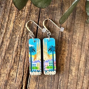 May include: A pair of silver dangle earrings with rectangular blue and white charms featuring a colorful illustration of Barcelona, Spain. The charms have the text "BARCELONA" printed on them.
