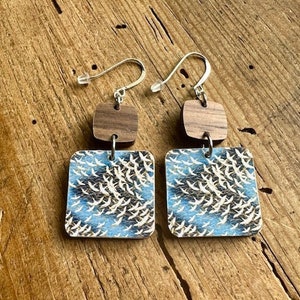 May include: A pair of wooden earrings with a blue and white bird print design. The earrings have a brown wooden square shape with a silver hook.