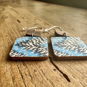 May include: A pair of square wooden earrings with a blue and white pattern of birds in flight. The earrings have silver hooks.