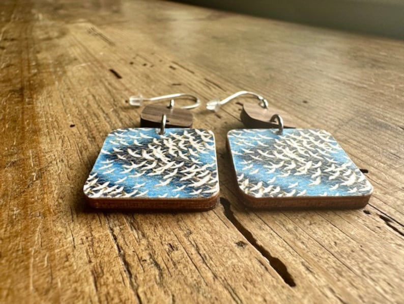 May include: A pair of square wooden earrings with a blue and white pattern of birds in flight. The earrings have silver hooks.