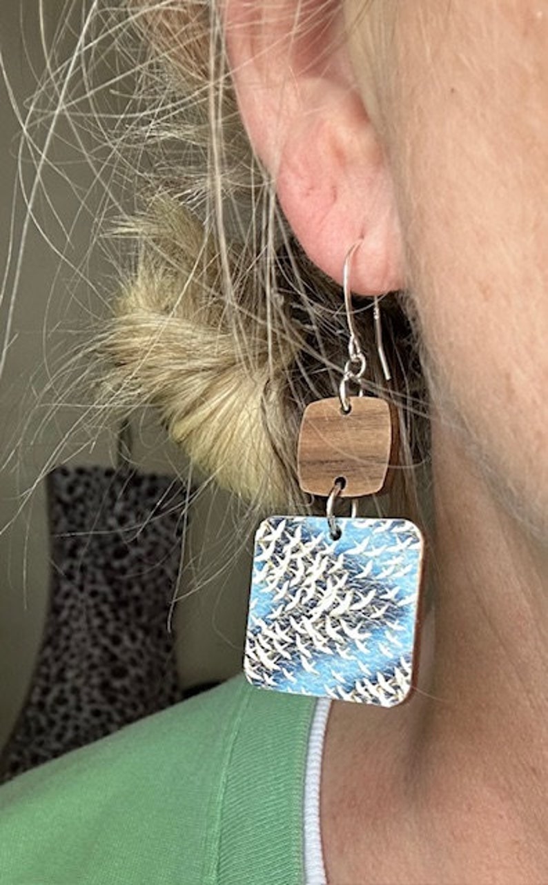 May include: A pair of dangle earrings with a square wooden top and a square bottom featuring a blue and white pattern of birds in flight.