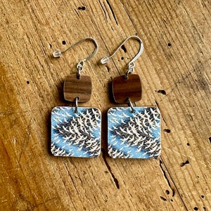 May include: A pair of wooden earrings with a blue and white pattern of birds in flight. The earrings are square shaped and have a brown wooden accent piece above the patterned piece.