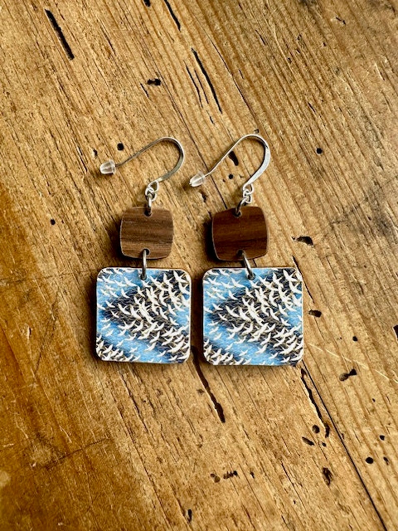 May include: A pair of wooden earrings with a blue and white pattern of birds in flight. The earrings are square shaped and have a brown wooden accent piece above the patterned piece.
