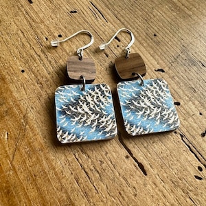 May include: A pair of wooden earrings with a blue and white pattern of birds. The earrings are made of two pieces, a square piece with the bird pattern and a smaller square piece of wood in a darker brown color. The earrings are attached to silver hooks.