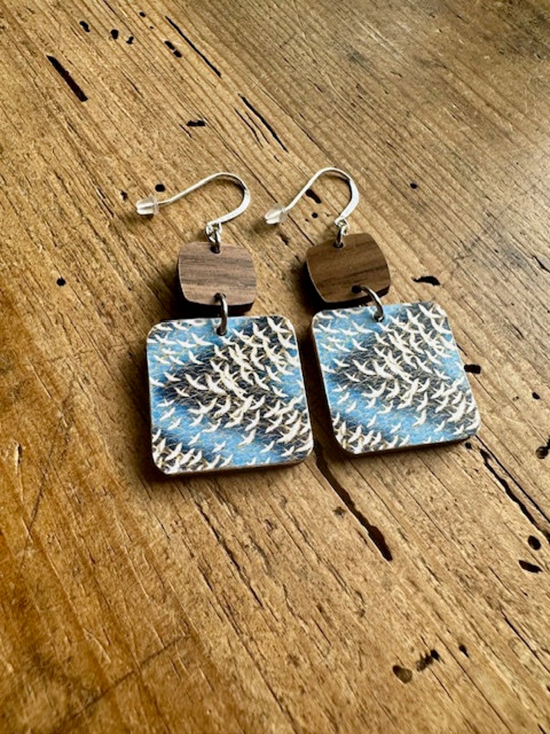 May include: A pair of wooden earrings with a blue and white pattern of birds. The earrings are made of two pieces, a square piece with the bird pattern and a smaller square piece of wood in a darker brown color. The earrings are attached to silver hooks.