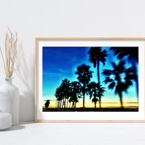 May include: A framed print featuring a row of silhouetted palm trees against a vibrant blue and orange sky. The image has a blurred effect, suggesting motion or a dreamy quality. The artwork is displayed on a white surface.