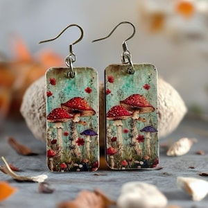 May include: A pair of rectangular earrings featuring a whimsical mushroom design. The earrings showcase red-capped mushrooms with white spots, set against a backdrop of flowers and foliage. The earrings are suspended from silver-colored ear wires.