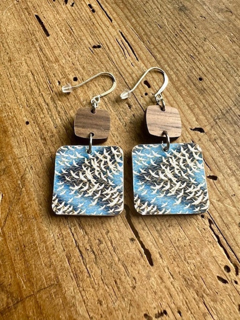 May include: A pair of wooden earrings with a blue and white bird print design. The earrings have a brown wooden square shape with a silver hook.