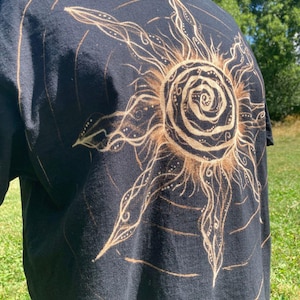 Puede incluir: Una camiseta negra de manga corta con un gran diseño de sol decolorado en la parte delantera. El gráfico del sol tiene un núcleo en espiral central con rayos radiantes, ondulados y puntiagudos decorados con pequeños puntos y líneas.
