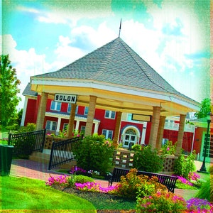 May include: A red brick building with a gazebo in front, featuring a sign that reads "SOLON." The gazebo has a gray roof and is supported by beige columns. The scene is framed by green grass, colorful flowers, and a blue sky.