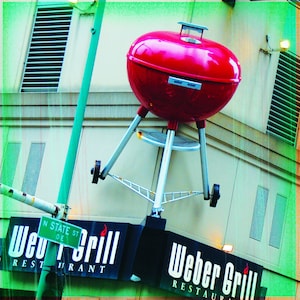 May include: A red Weber grill with a round shape, supported by silver legs and wheels. The image also shows a sign that says "Weber Grill Restaurant" and a street sign that says "N State St".