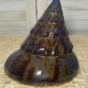 Puede incluir: Una escultura de cerámica cónica con un esmalte brillante que presenta vetas mezcladas de azul profundo y marrón oscuro. El objeto tiene una estructura en espiral de varios niveles que se estrecha hacia la punta, con una superficie lisa.