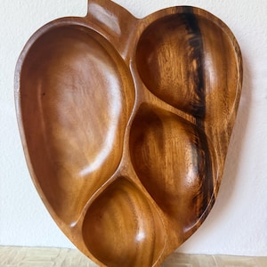 May include: A polished wooden serving tray shaped like an apple with a stem at the top. The tray features four distinct, carved compartments of varying sizes, showcasing a natural brown wood grain pattern with darker streaks throughout the surface.