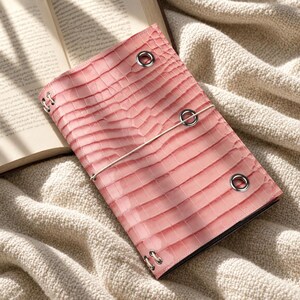 May include: A rectangular notebook cover in a light pink color with a textured crocodile skin pattern. The cover features four silver-toned metal eyelets at the corners and a central elastic band closure that wraps horizontally around the exterior.