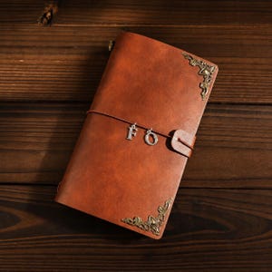 May include: A rectangular brown notebook with a smooth cover, featuring ornate brass-coloured metal corner protectors on the top right and bottom left. A thin cord wraps around the centre, holding two silver-toned letter charms, F and O.