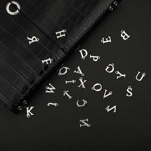 May include: A collection of silver-toned metal alphabet letter charms scattered across a dark surface. Each individual letter charm features a small loop at the top for attachment, showcasing a variety of uppercase characters in a classic serif font.