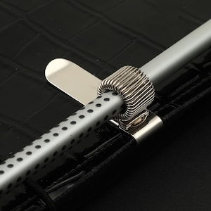 May include: A silver metal pen holder clip attached to a black crocodile-textured surface. The clip features a coiled spring loop holding a light gray pen with a black polka dot pattern. The clip has a flat, smooth, reflective metal tab extension.