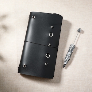 May include: A black leather journal cover featuring several silver-toned metal eyelets of varying sizes scattered across the front surface, secured by a thin black elastic cord with a small star charm, accompanied by a silver textured ballpoint pen.