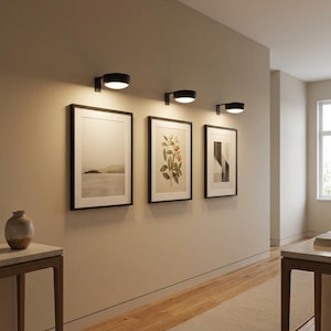 May include: Three black wall-mounted sconces with circular heads provide downward illumination over three framed rectangular wall art pieces. The artwork includes a landscape, a botanical illustration, and an abstract geometric composition.