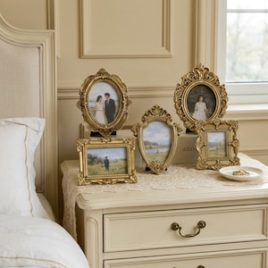May include: A collection of four ornate gold-toned picture frames in various shapes, including oval, rectangular, and shield-like designs. Each frame features intricate embossed detailing along the edges and holds a photograph or landscape image.