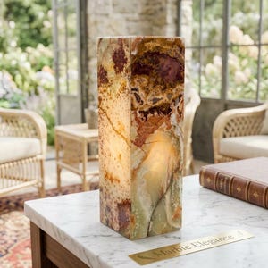 May include: A rectangular onyx stone lamp featuring a polished surface with natural veining in shades of amber, brown, cream, and translucent green. The lamp emits a soft internal glow and sits on a white marble table next to a gold plaque.