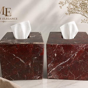 Pode incluir: Duas caixas quadradas para lenços feitas de mármore vermelho escuro com veios brancos proeminentes. Cada caixa apresenta uma abertura circular central no topo com um lenço branco saliente. O logótipo ME MARBLE ELEGANCE é visível no canto superior esquerdo.