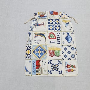 May include: A cream-coloured drawstring pouch featuring a grid pattern of colourful illustrations including blue and yellow tile motifs, a boat, a fish, striped beach huts, and the text I LOVE AVEIRO, along with small figures in traditional clothing.
