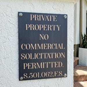 May include: A rectangular black sign with light-colored lettering that reads PRIVATE PROPERTY NO COMMERCIAL SOLICITATION PERMITTED. S.501.062,F.S. The sign is mounted with four silver-toned metal screws at each corner on a textured white wall.