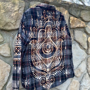 May include: A grey, black, and red checked flannel shirt with a detailed tan design on the back. The design includes a hand with an eye, surrounded by flame-like shapes and other decorative elements, creating a unique aesthetic.