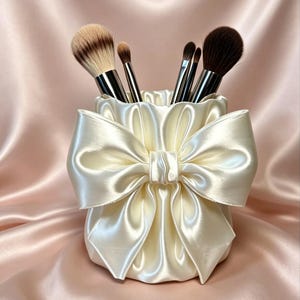 Peut inclure: Un porte-pinceaux de maquillage cylindrique en satin couleur crème, orné d'un grand nœud plissé en satin sur le devant. Le support contient quatre pinceaux de maquillage avec des viroles argentées et des poils dans des tons beige, brun et noir.