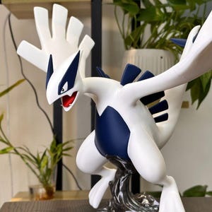 Puede incluir: Una figura de Lugia blanca y azul oscuro con una gran envergadura, cuello largo y boca roja. La criatura está en una postura dinámica, apoyada sobre una base texturizada de color gris oscuro que se asemeja a agua o humo arremolinado.