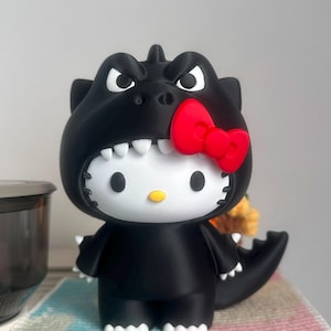 Puede incluir: Una figura de vinilo de un personaje de gato blanco que lleva un disfraz de monstruo negro con capucha. El disfraz presenta un lazo rojo en el lateral, dientes blancos a lo largo de la abertura de la capucha y una cola.
