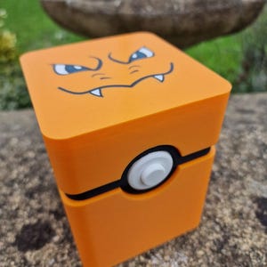May include: A square orange storage box featuring a stylised graphic of a creature face with sharp teeth on the top lid. The box has a black horizontal band wrapping around the centre with a circular white button centred on the front face.