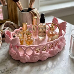 Puede incluir: Una bandeja de tocador ovalada de color rosa claro con un borde estilo celosía y lazos esculpidos en los laterales. La bandeja contiene cuatro frascos de perfume de vidrio de diversas formas y tamaños, cada uno con tapa dorada, y un atomizador.