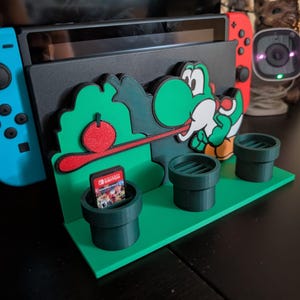 May include: A black Nintendo Switch console dock featuring a green Yoshi character graphic, a red fruit accent, and three dark green cylindrical game cartridge holders on a flat base, with a blue and red controller attached to the console.