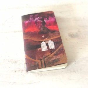 May include: A small, brown notebook with a colorful abstract design on the cover. The design features pink and red flowers, a brown background, and white paper scraps with text.