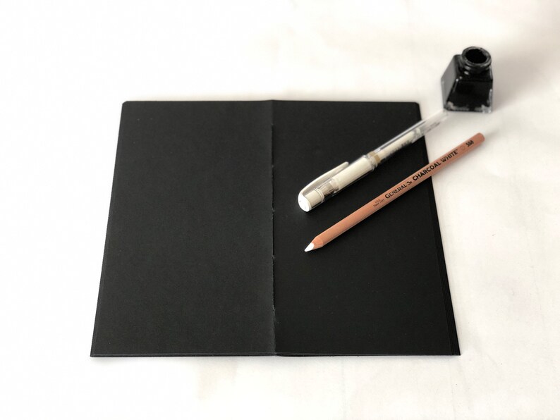 May include: A black sketchbook with a white pen and a brown pencil with the text "GENERAL'S CHARCOAL WHITE 308" on it.