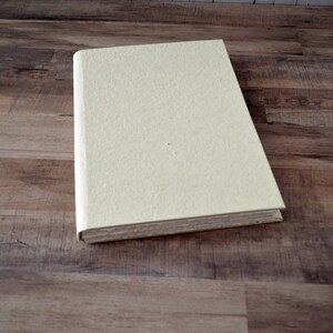 May include: A cream-colored, blank journal with a textured cover. The journal is closed and lying flat on a wood surface.