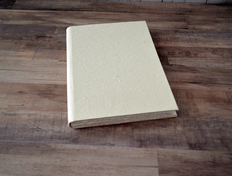 May include: A cream-colored, blank journal with a textured cover. The journal is closed and lying flat on a wood surface.