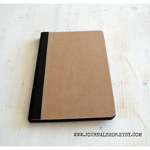 Pode incluir: Um caderno marrom com uma lombada preta. O caderno est&aacute; fechado e deitado sobre uma superf&iacute;cie de madeira branca.