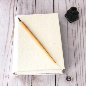 May include: A white blank journal with a wooden calligraphy pen and a small black inkwell on a light wood surface.