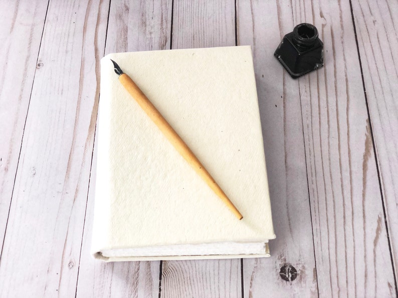 May include: A white blank journal with a wooden calligraphy pen and a small black inkwell on a light wood surface.