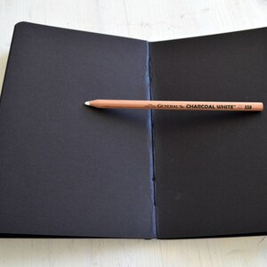 Pode incluir: Um caderno de desenho preto com um l&aacute;pis em cima. O l&aacute;pis est&aacute; etiquetado como "GENERAL'S CHARCOAL WHITE 530".