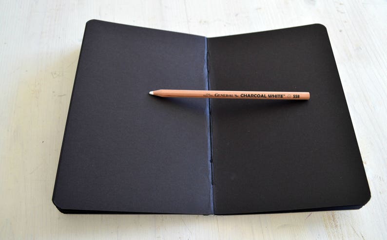 Pode incluir: Um caderno de desenho preto com um l&aacute;pis em cima. O l&aacute;pis est&aacute; etiquetado como "GENERAL'S CHARCOAL WHITE 530".