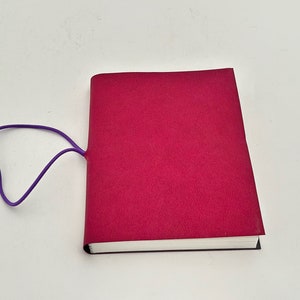May include: A burgundy notebook with a purple elastic band closure. The notebook is closed and lying flat on a white surface.