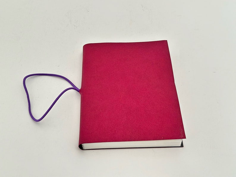May include: A burgundy notebook with a purple elastic band closure. The notebook is closed and lying flat on a white surface.