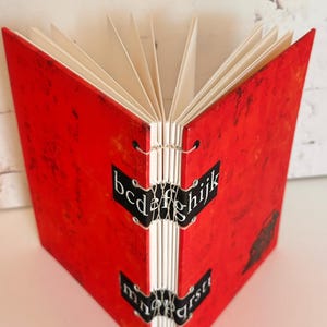 Puede incluir: Diario rojo hecho a mano con páginas blancas y letras negras en el lomo que dicen "bcd efg hijk mn opq rst".